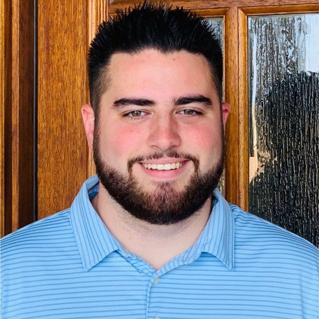 A man with a beard and a blue shirt is smiling in front of a wooden door.