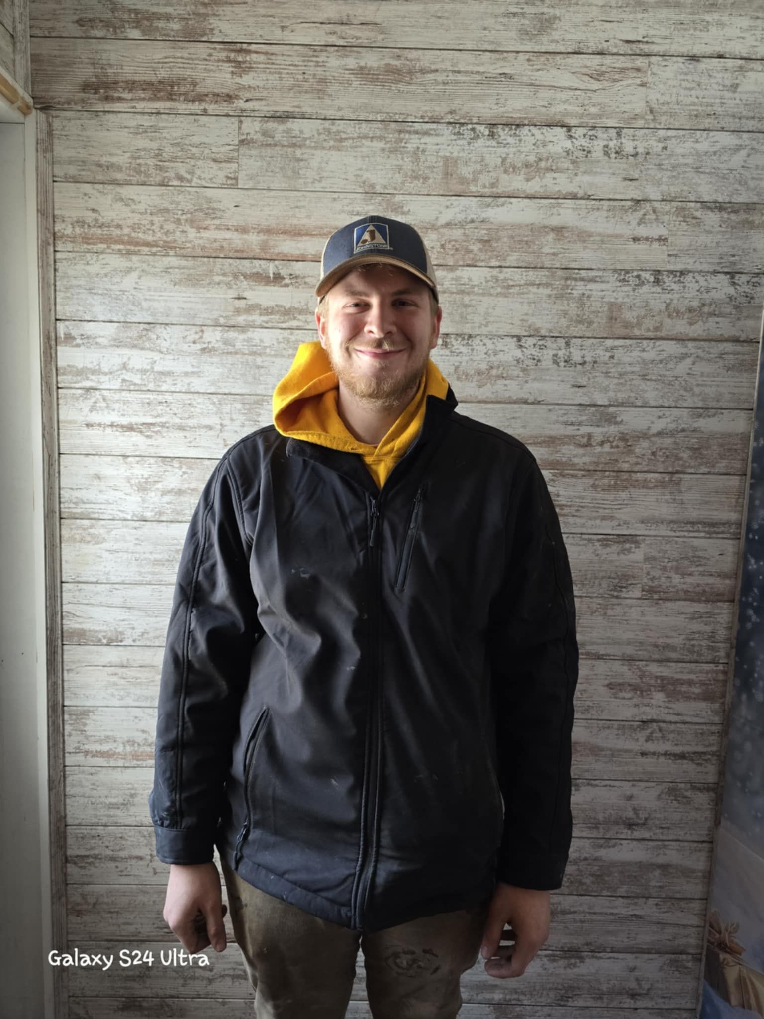 A man wearing a black jacket and a hat is standing in front of a wooden wall.
