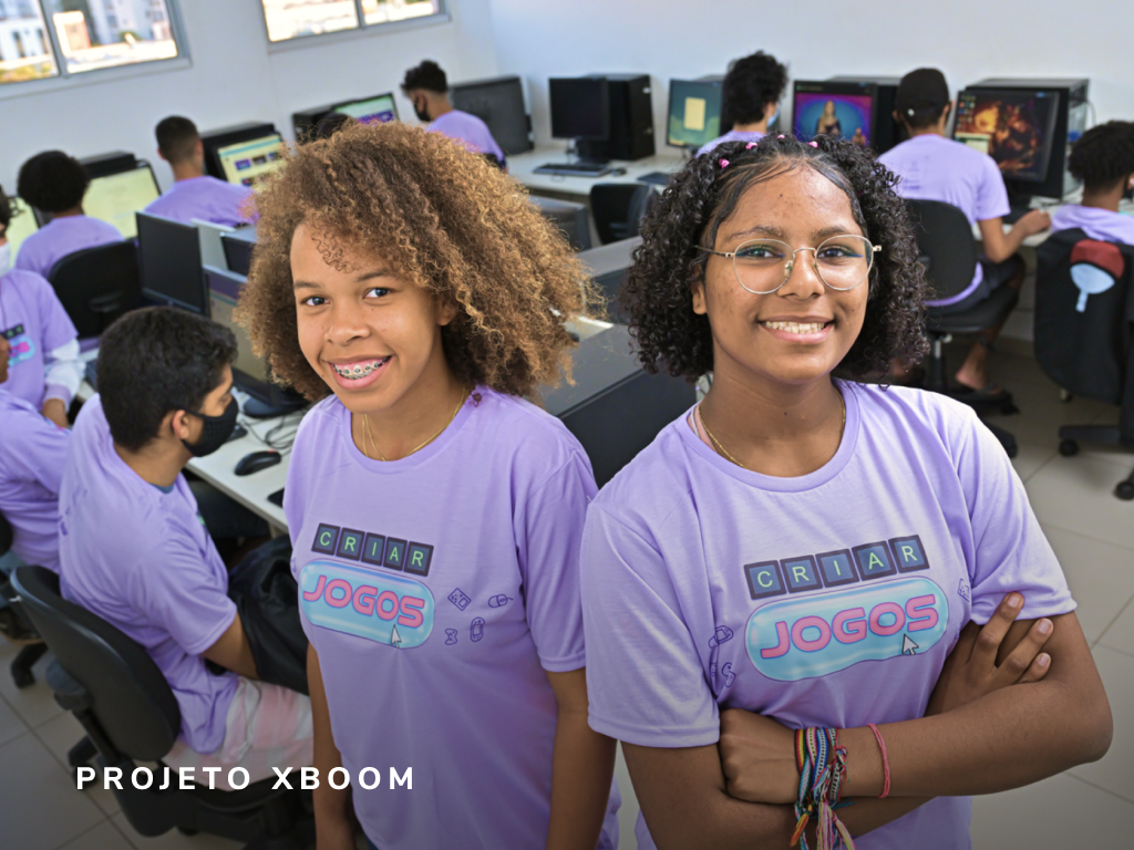Duas garotas de blusa roxa estão posando para uma foto em um laboratório de informática.