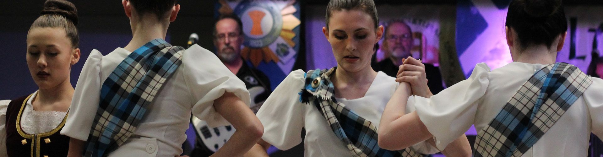 Two dancers in blue plaid costumes hold hands in a dimly lit stage performance