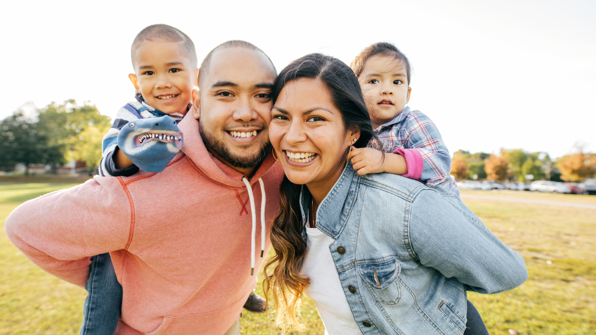 Family smiling outside. Both dad and mom have a child riding piggyback on their backs. 