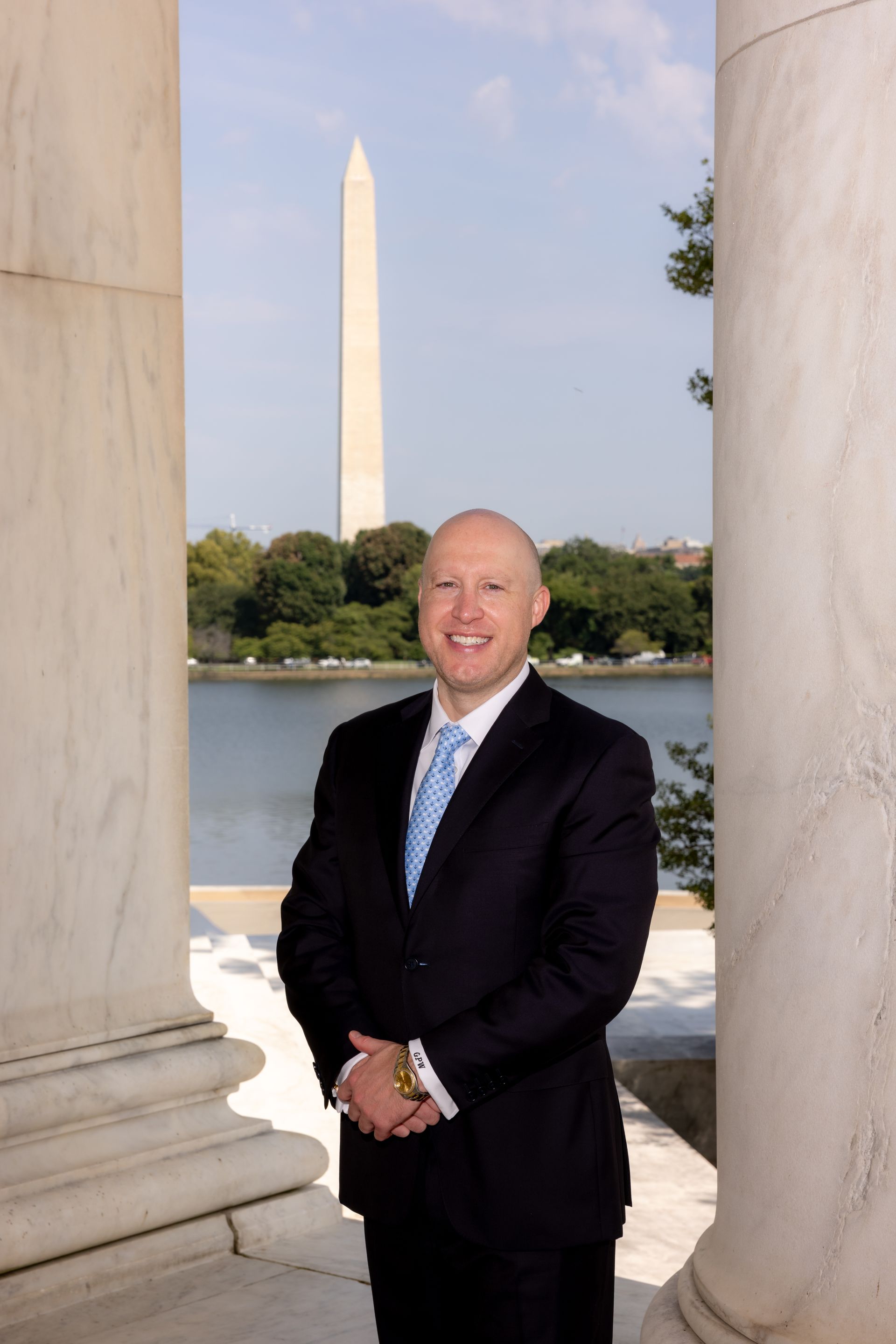 Man in a dark suit standing between white columns with the Washington Monument and reflecting pool behind him