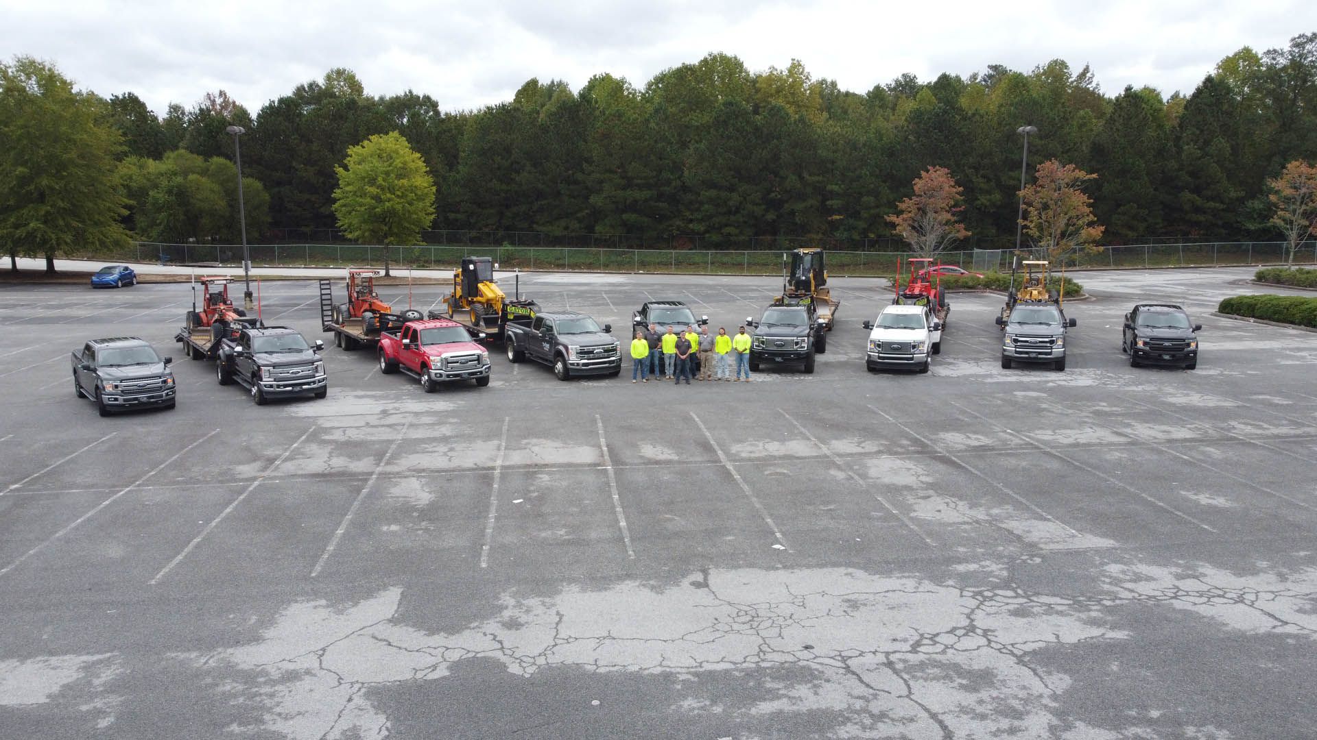Group of construction workers and vehicles in a parking lot; trees in the background.