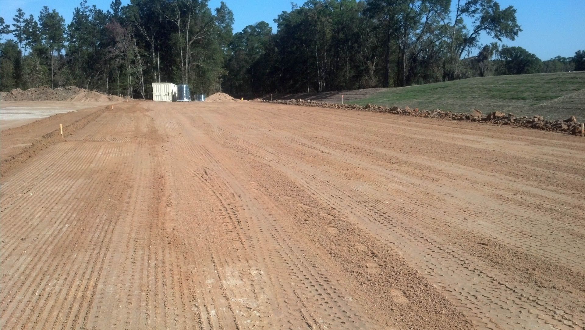Dirt road under construction, with tire tracks. Trees and a small building are visible in the background.