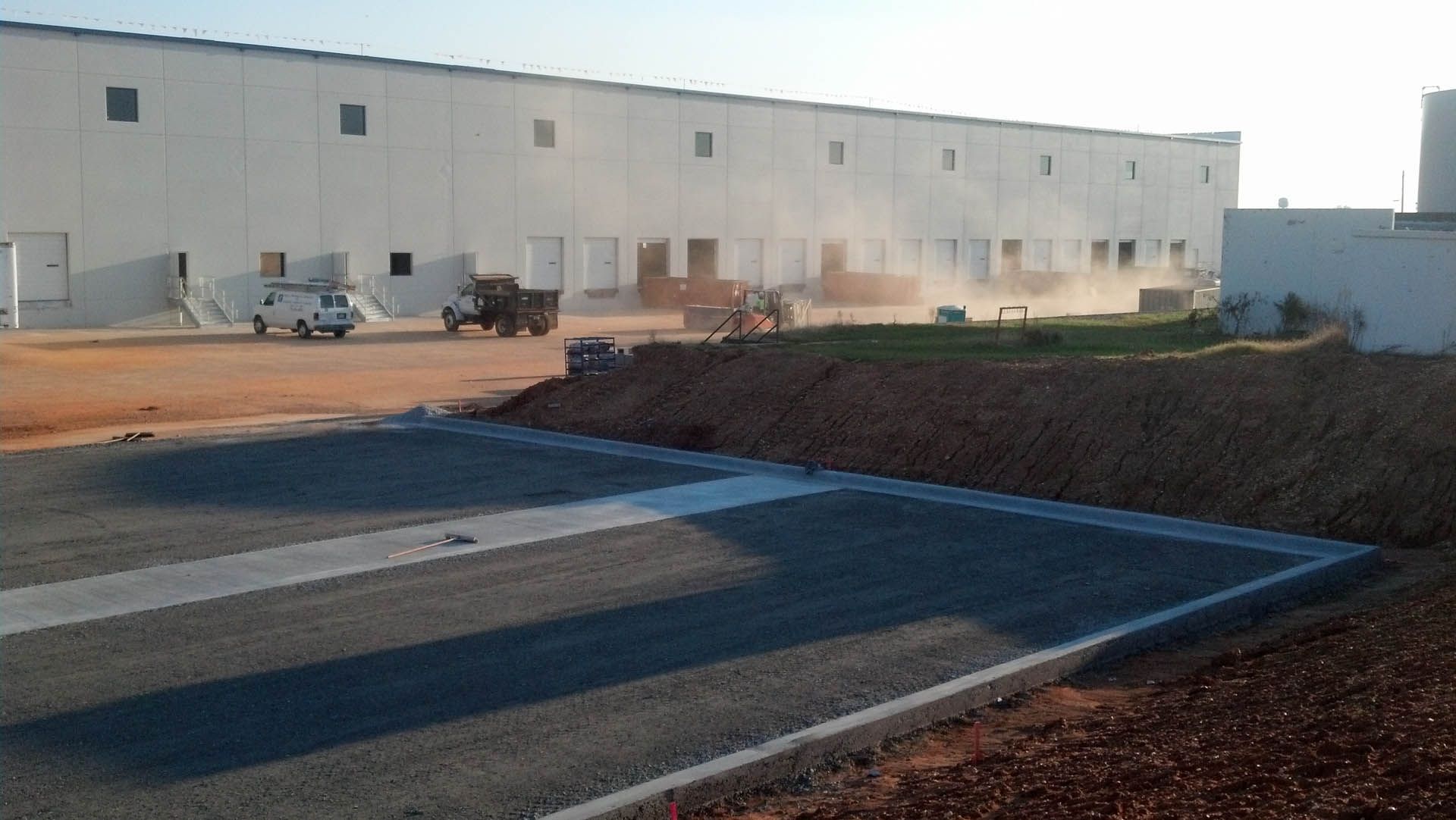 Construction site with a long, white warehouse. Workers and vehicles in operation. Gravel and dirt in the foreground.