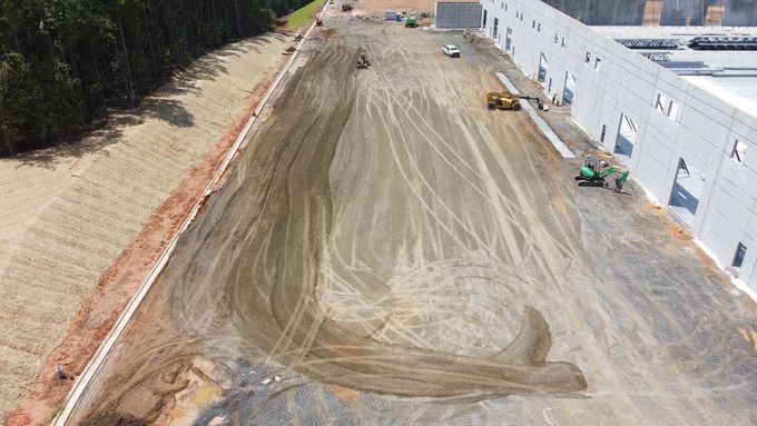 Construction site with dirt ground being graded next to a large building, trees, and heavy machinery.