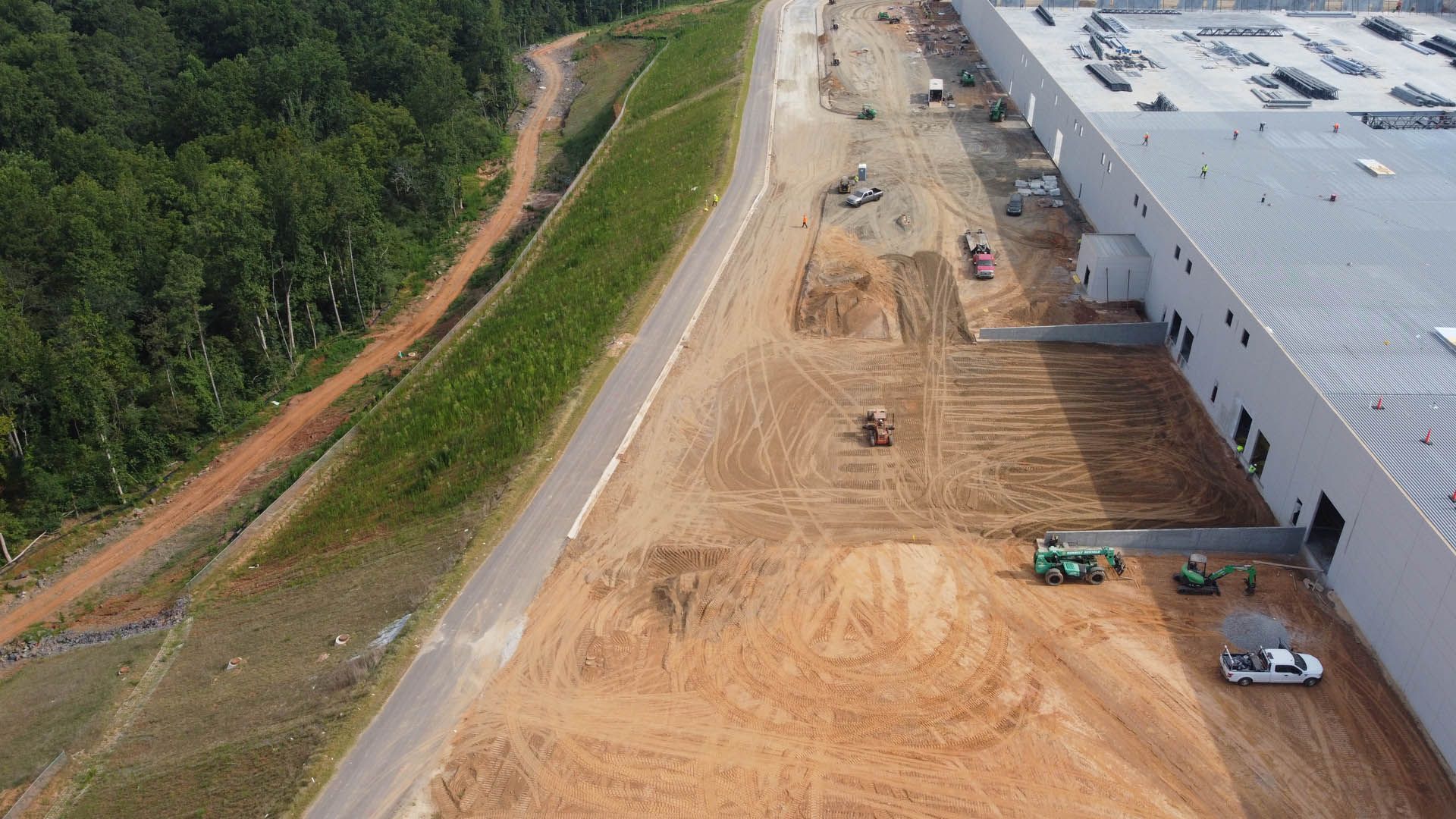 Aerial view of a construction site with a large building, dirt, and equipment near trees.