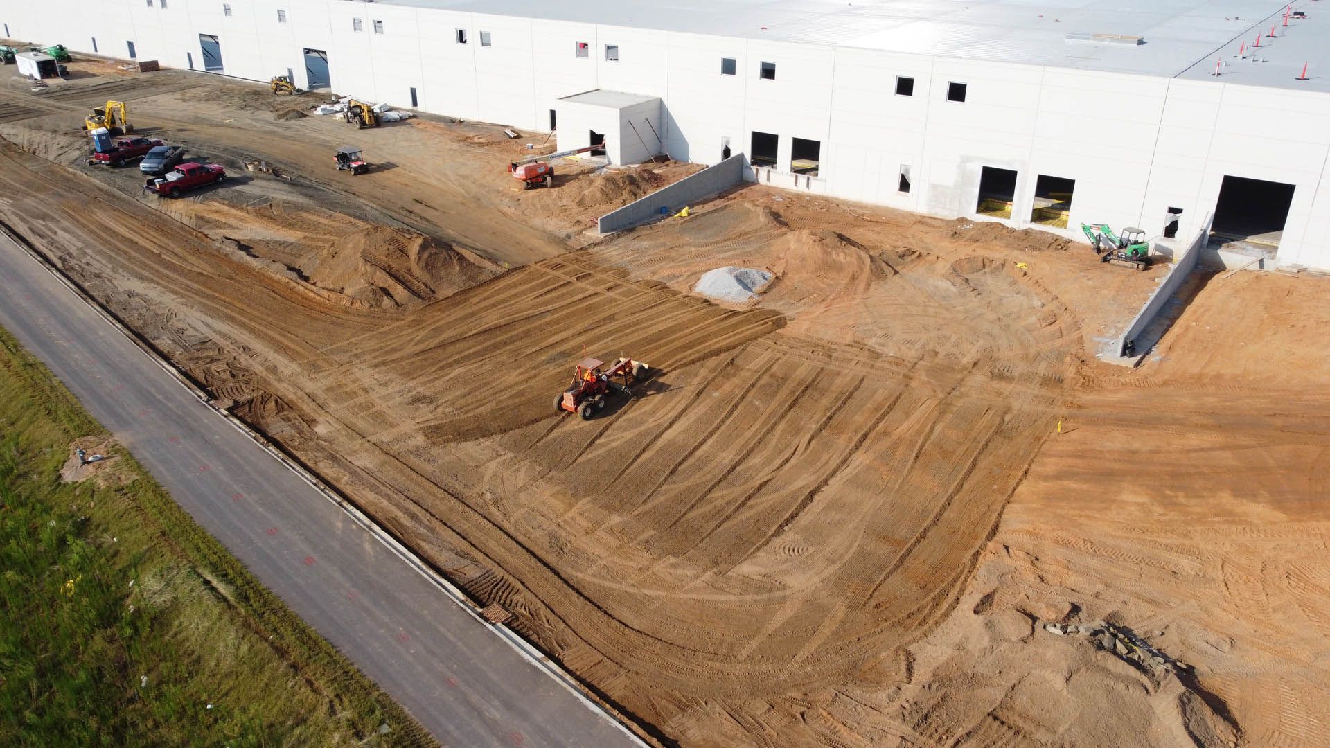 Construction site with heavy machinery grading the brown earth in front of a white industrial building.