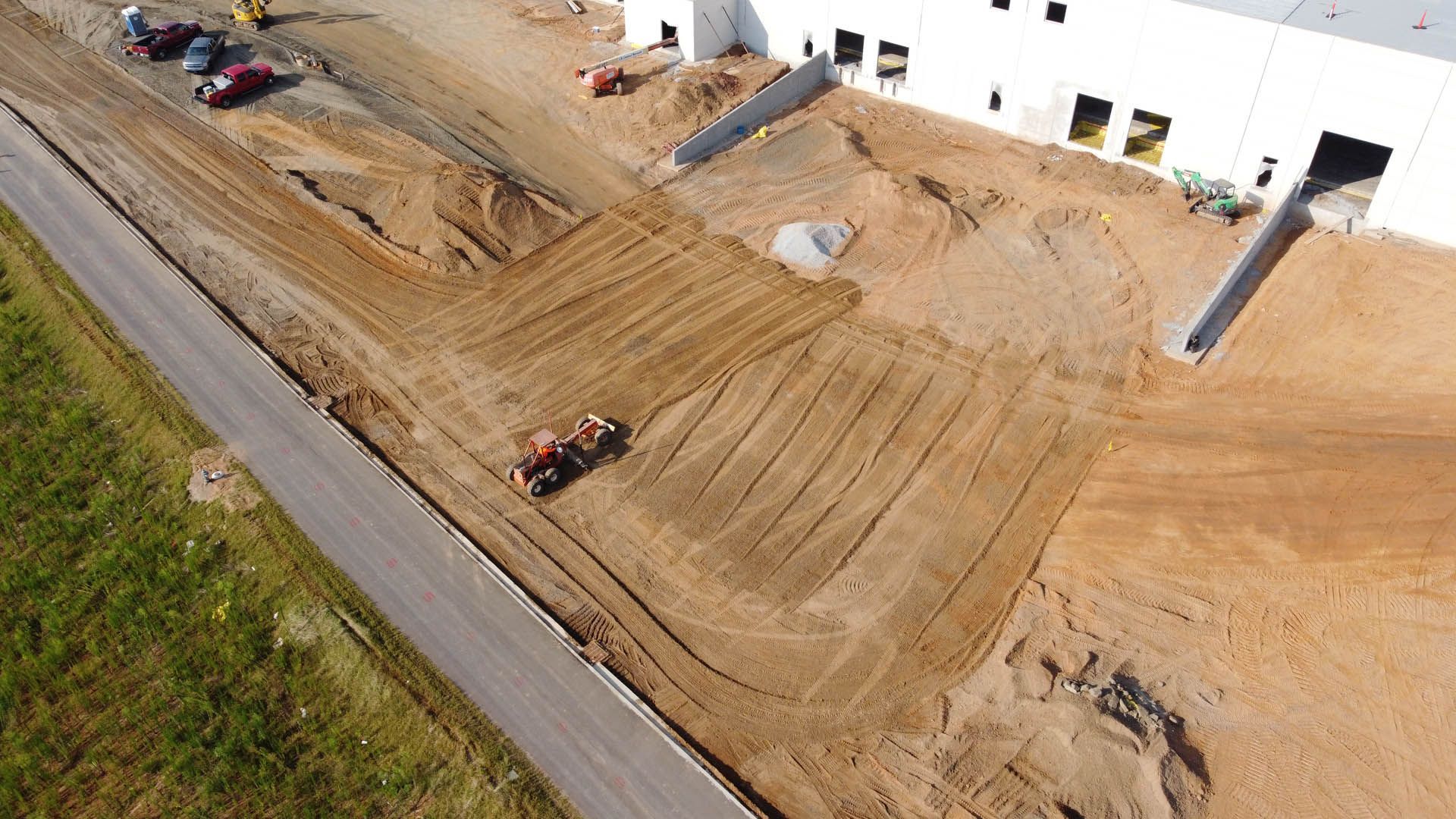 Construction site with building, tractor, and graded dirt. Asphalt road and green grass present.