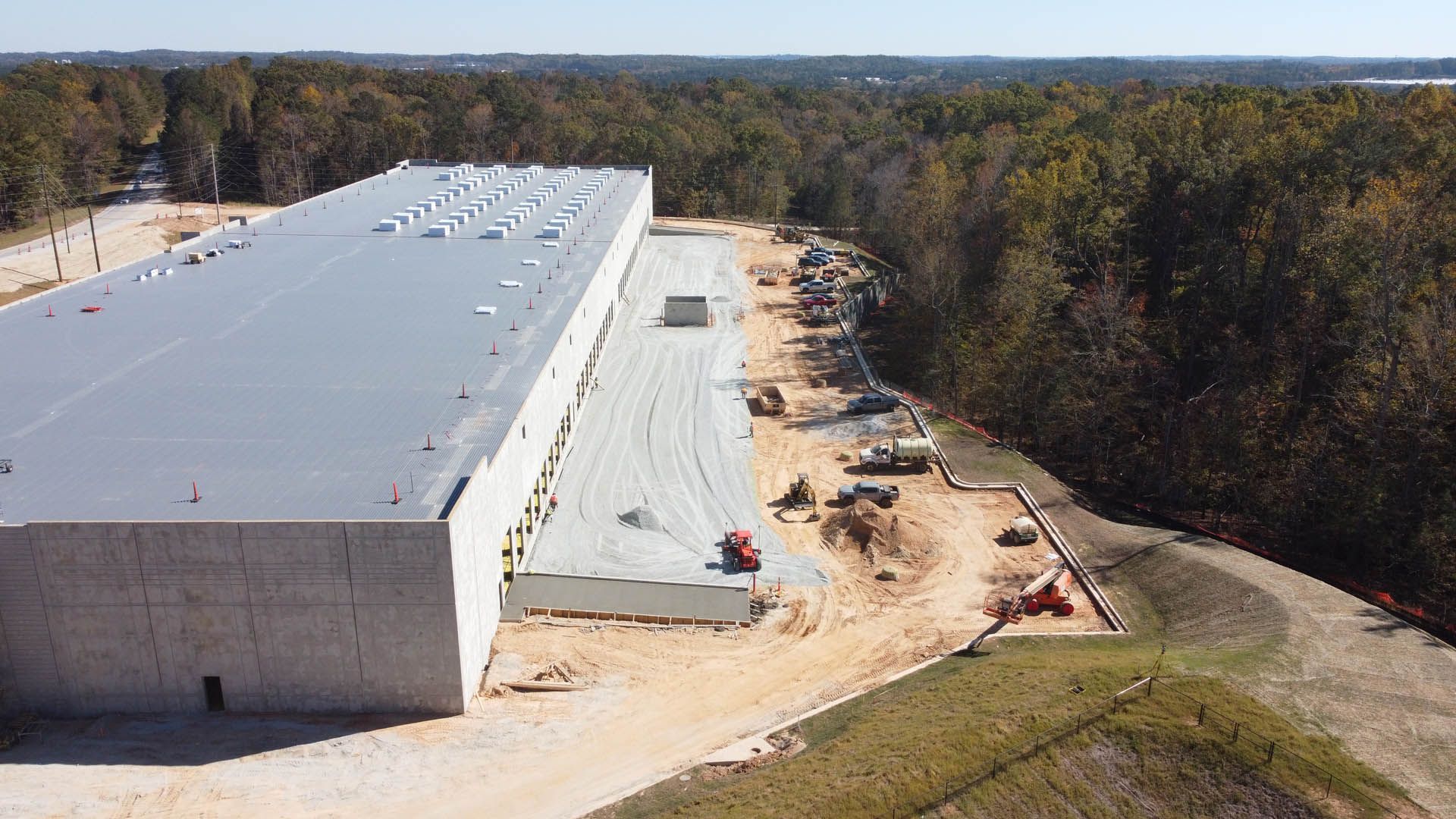 Large gray industrial building under construction, surrounded by dirt and trees, sunny day.