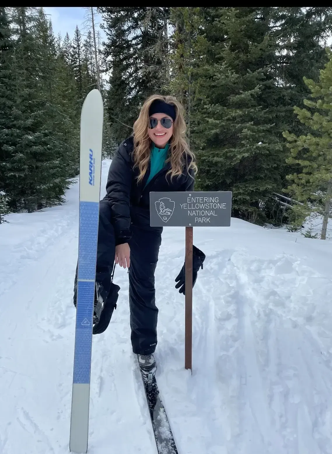 Person in black winter gear standing on snow beside skis and a trail sign in a snowy forest