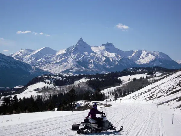 Snowmobile rider on a snowy trail with mountain peaks under a clear blue sky.
