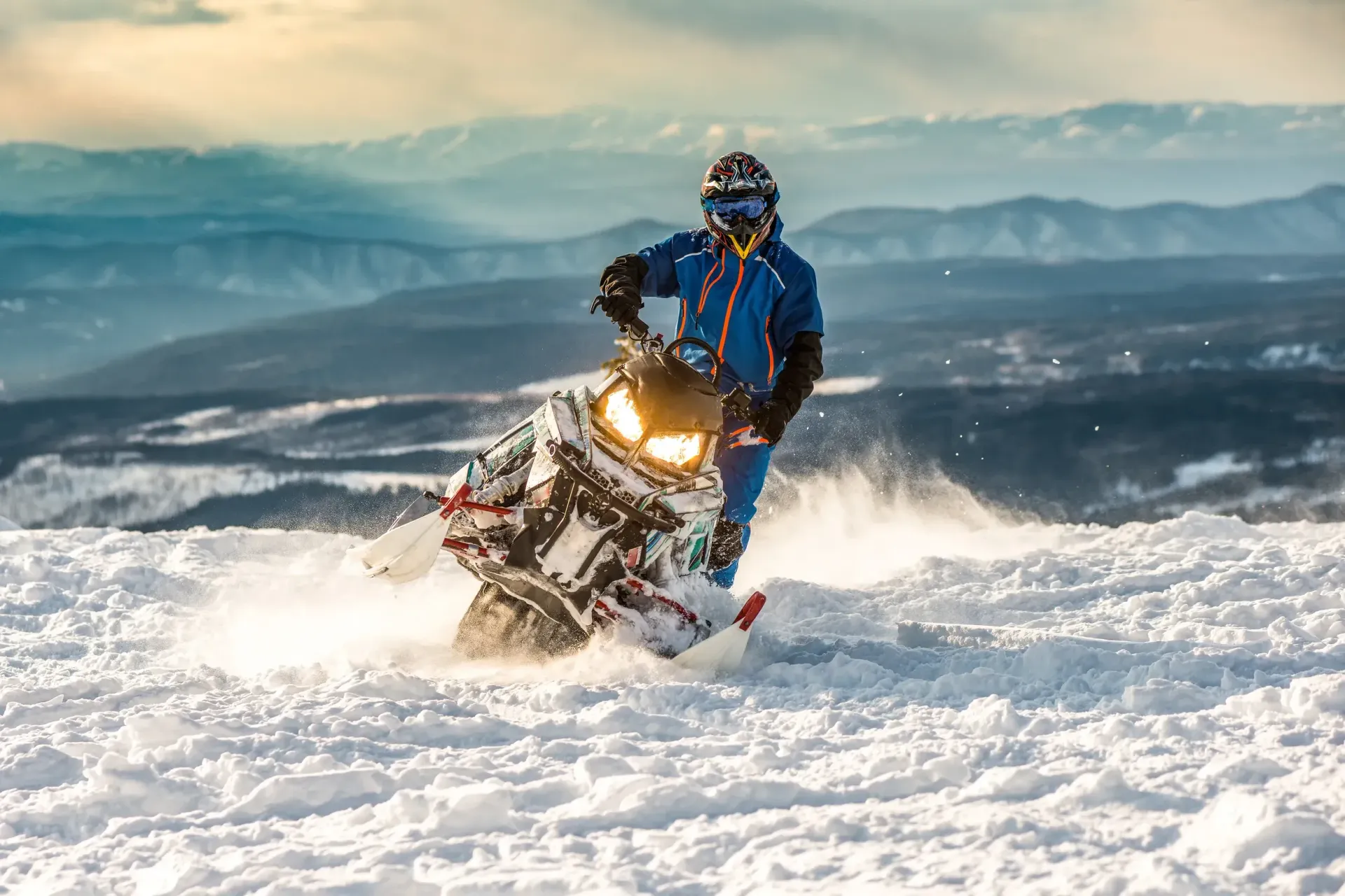 Snowmobiler in blue gear racing through fresh snow with mountains in the background