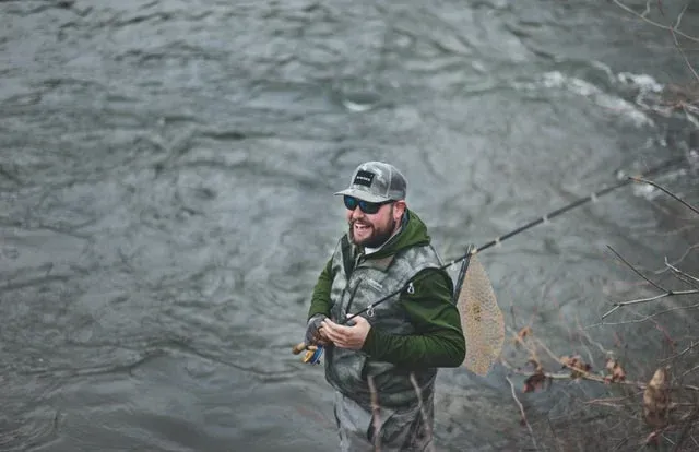 Angler in waders fishing beside a fast-moving river with a net on the bank
