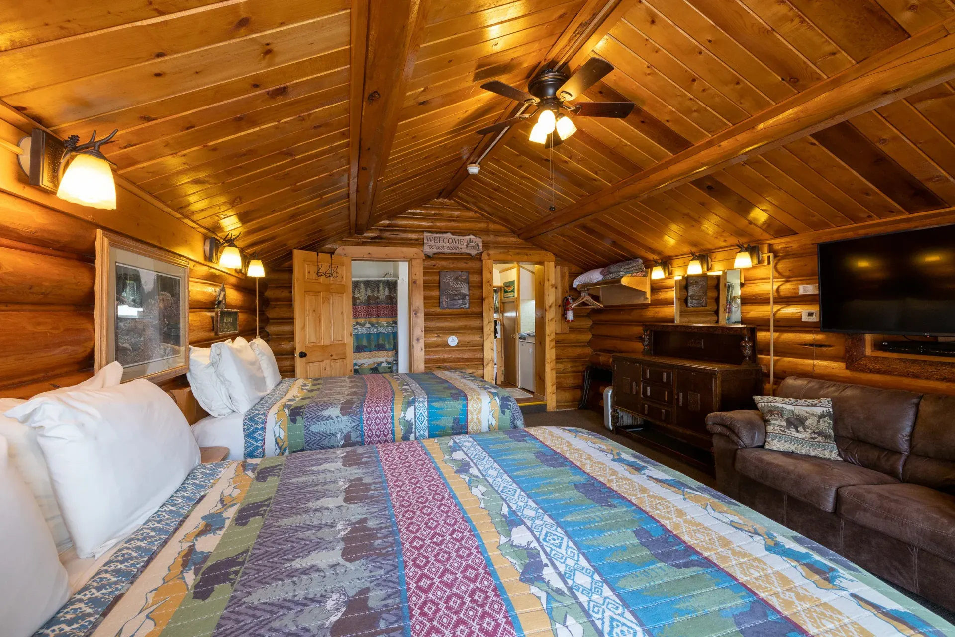 Cozy log cabin bedroom with a large colorful bedspread, wood-paneled ceiling, and TV on the wall