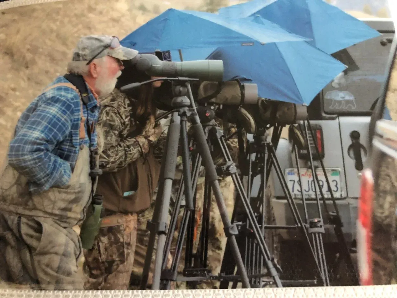 Two men on tripods with telescopes under blue umbrellas in rainy weather