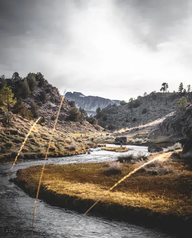 Mountain valley with a winding river, rocky hills, and golden grass under a cloudy sky