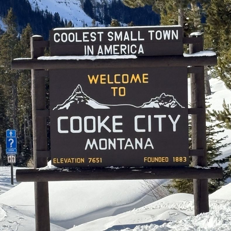 Snowy mountain road with a welcome sign for Eagle Lake, Montana, beside tall pines.