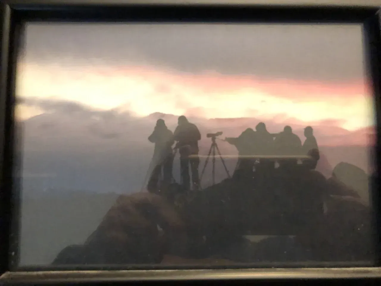 Silhouetted people on a mountain overlook at sunset, with a camera tripod and cloudy sky.