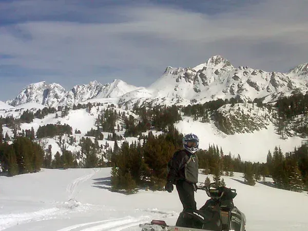 Snowmobile rider on a snowy trail with pine trees and rugged mountains in the background