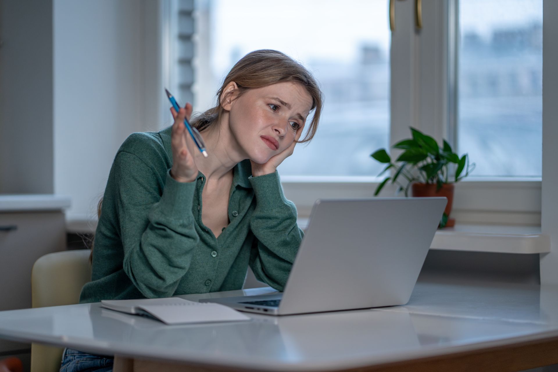 Woman at a desk using a laptop, holding a pen, with papers and a potted plant by a window.