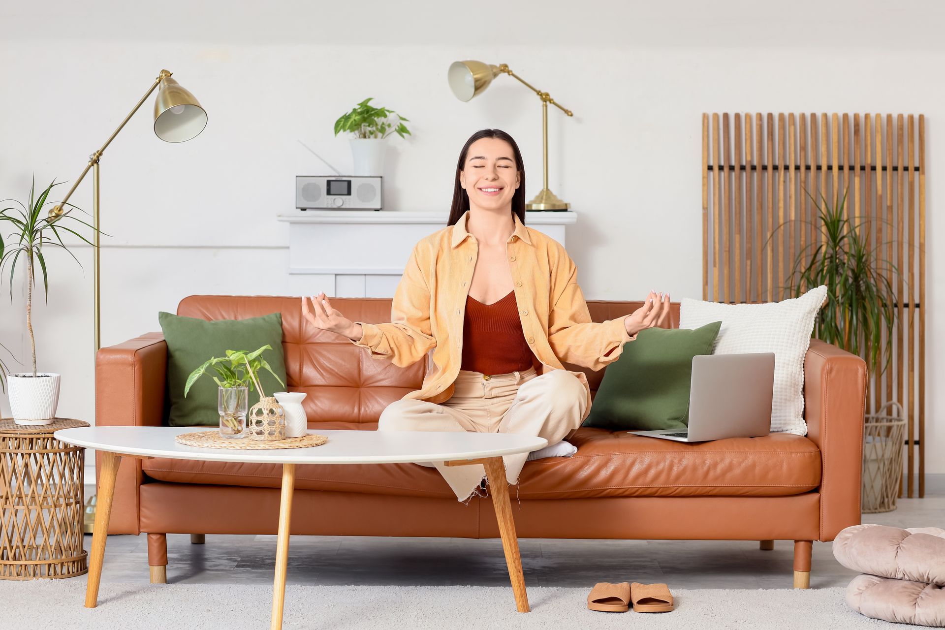 Woman meditating on a tan sofa in a bright living room with plants and warm decor