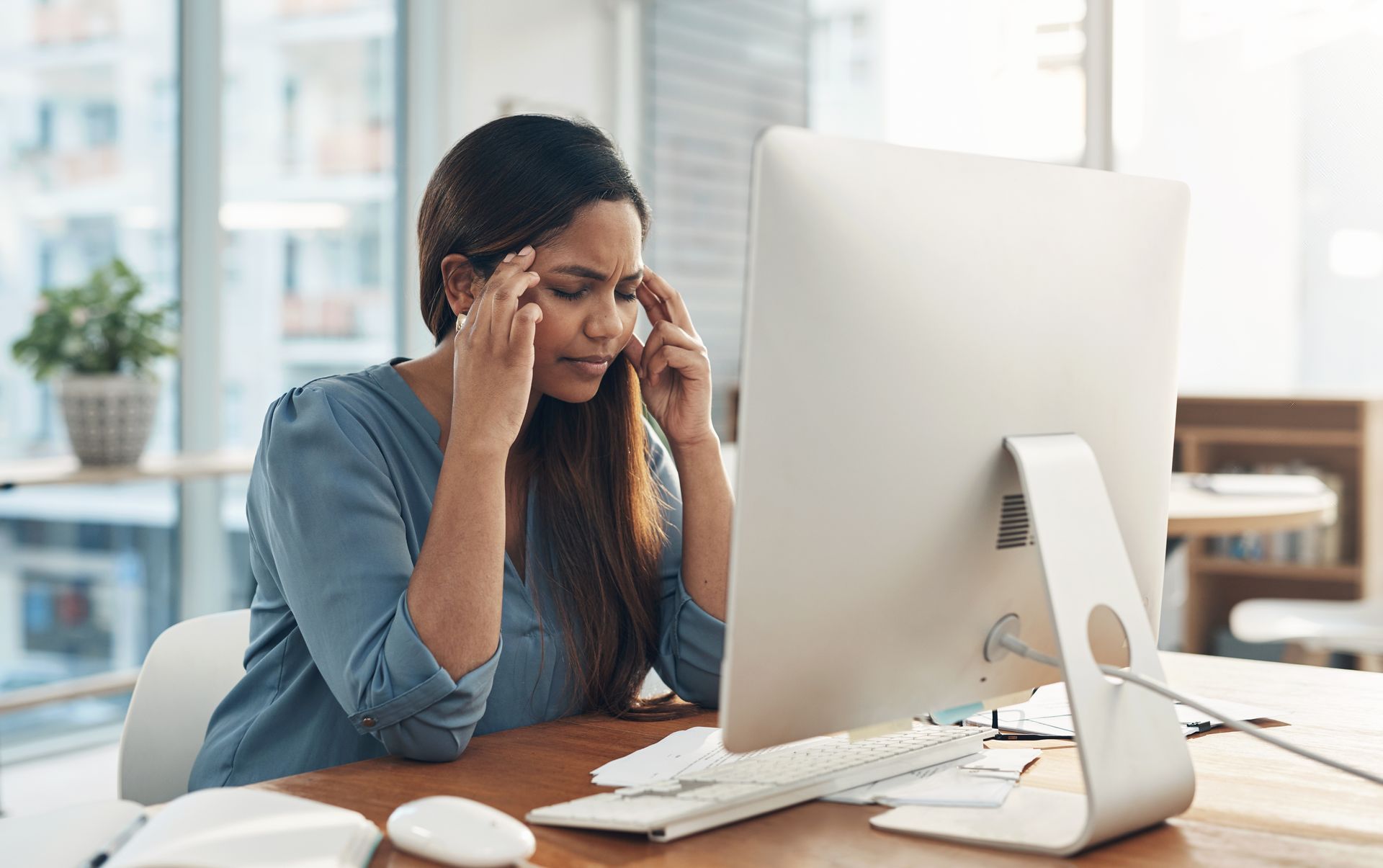 Woman rubbing her temples at a desk in front of a computer, looking stressed in a bright office