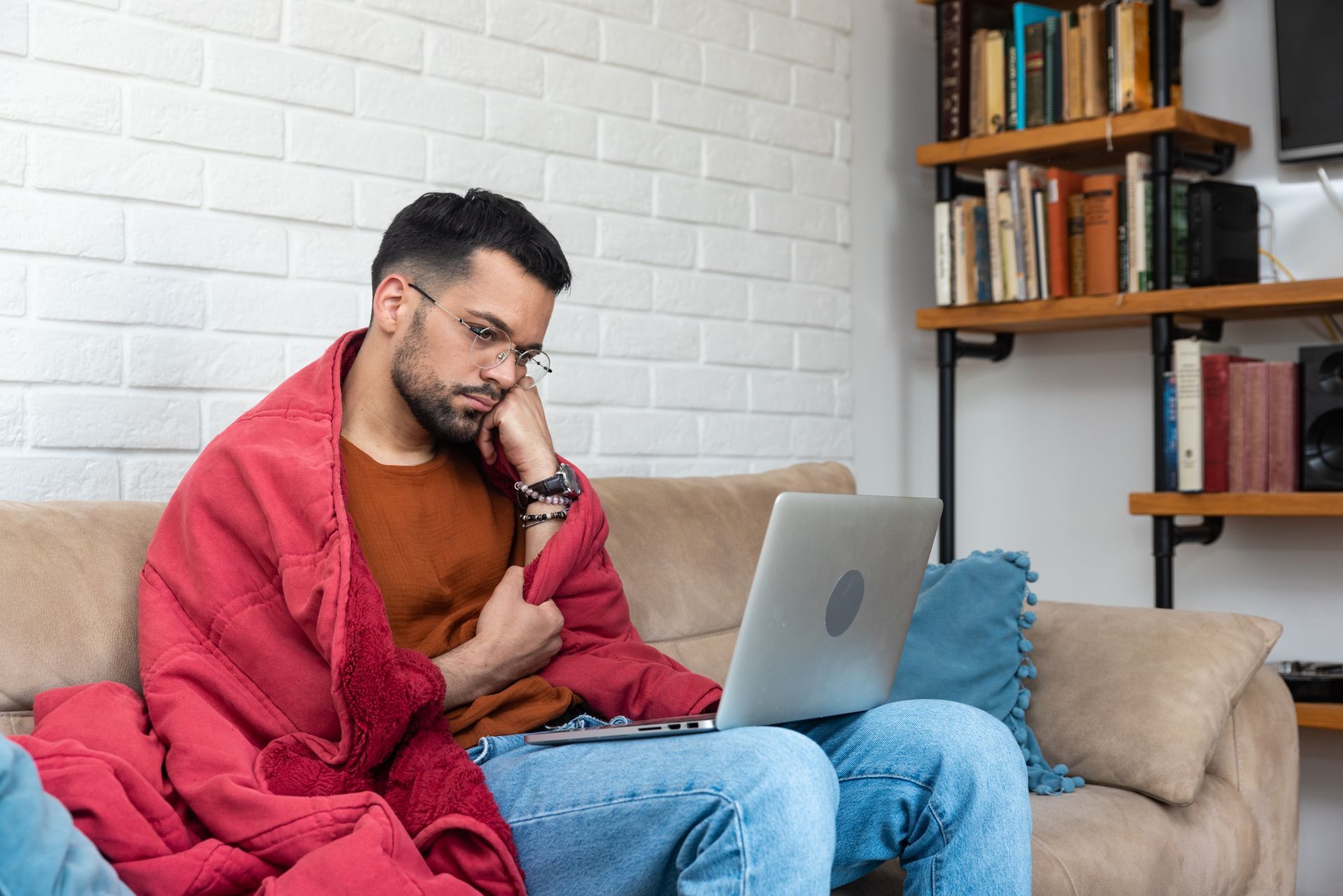 Man wrapped in a red blanket sits on a couch, talking on a phone beside a laptop in a cozy living room
