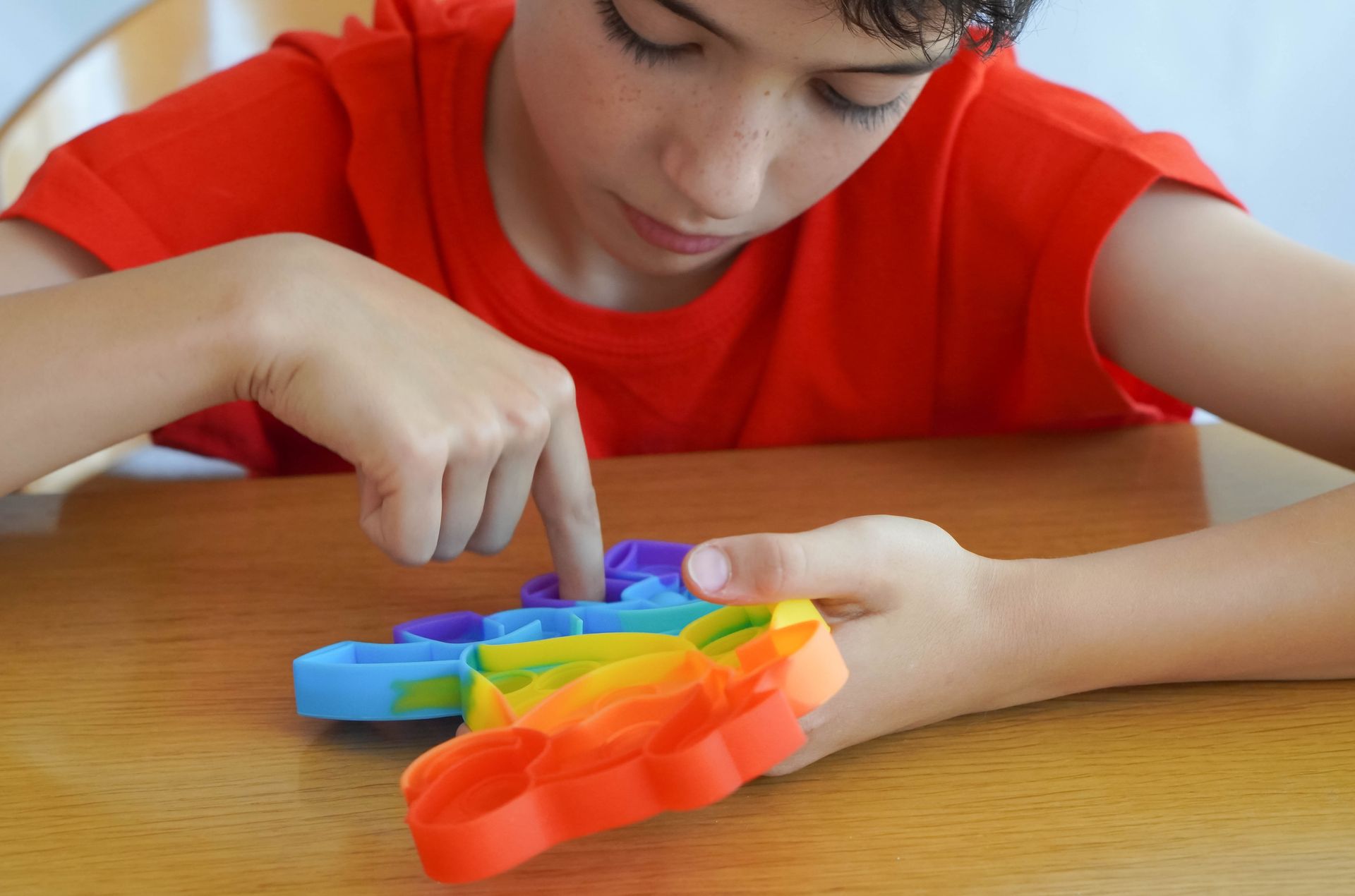 Child in a red shirt playing with a colorful rainbow pop-it toy on a table