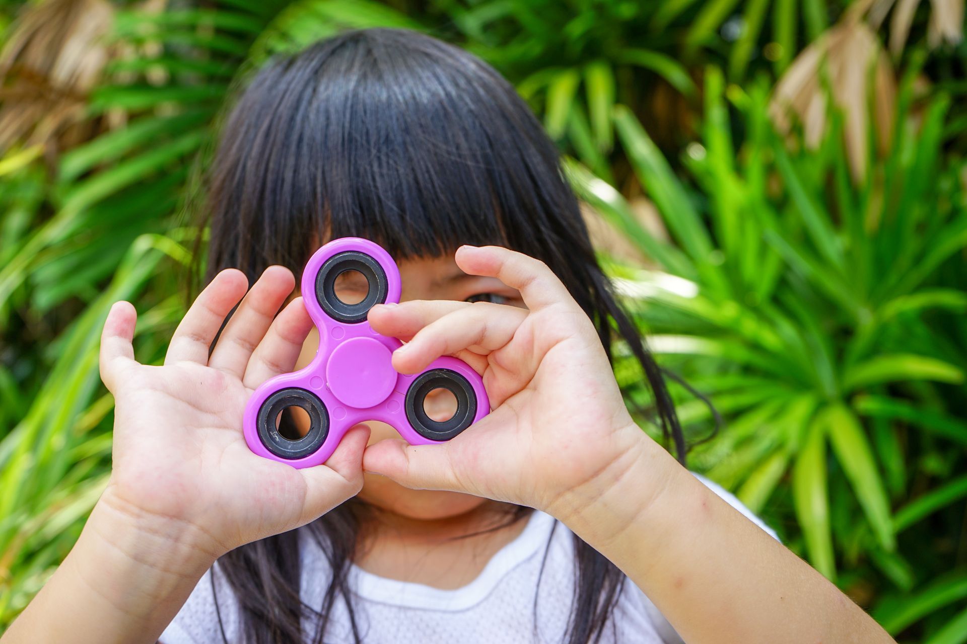Child holding a pink fidget spinner in front of their face with green plants in the background
