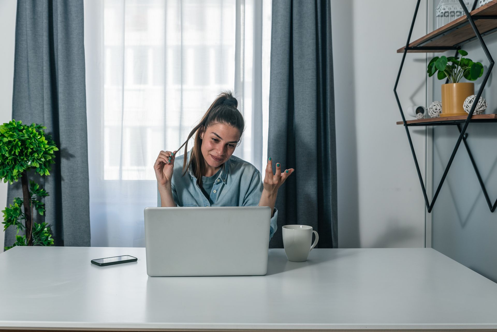 Woman using laptop at a white desk in a bright home office, with plants and shelves in the background.