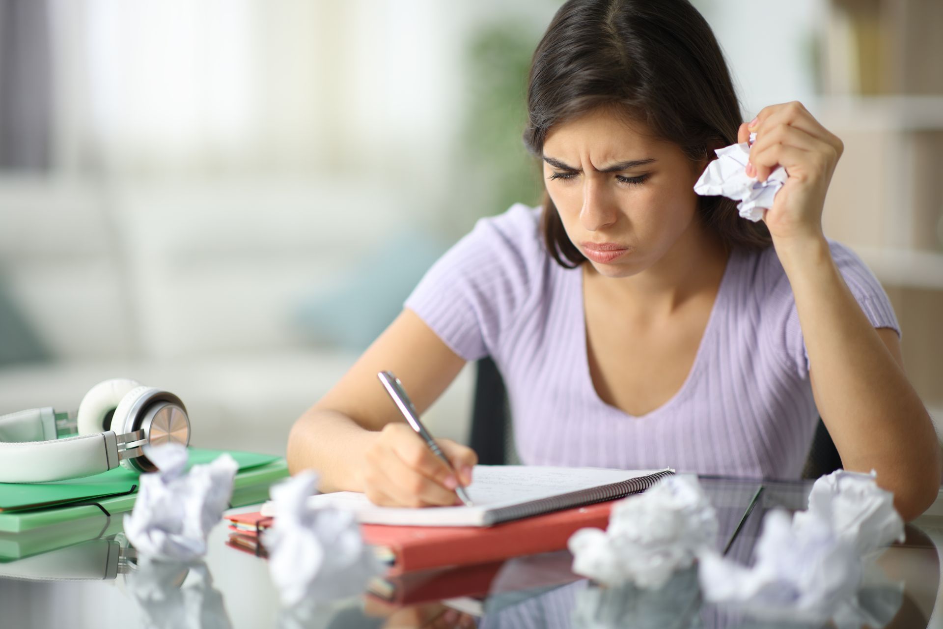 Frustrated student studying at a desk, holding tissue while writing amid crumpled papers