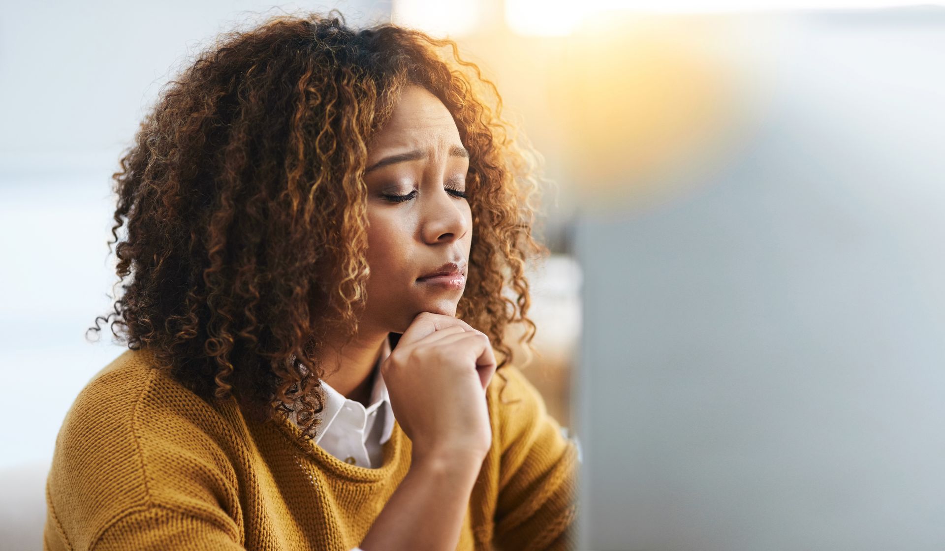 Person in a mustard sweater sits thoughtfully by a window, chin resting on hands.