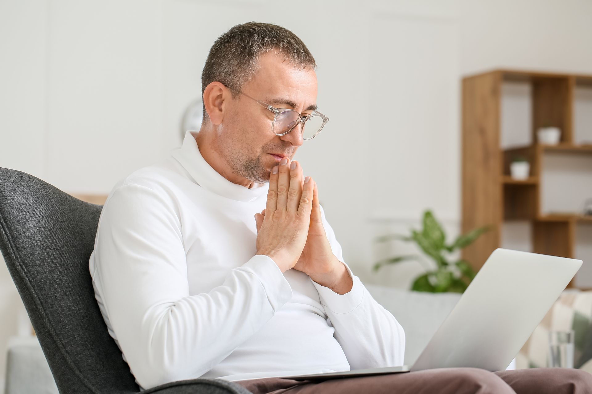 Man in glasses sitting on a couch, hands clasped in prayer beside an open laptop