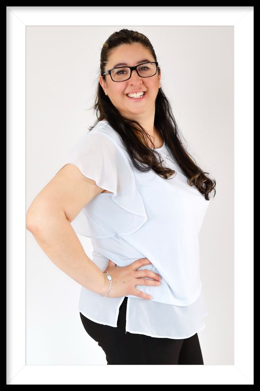 Woman with glasses smiling, hand on hip, wearing a light blue top and black pants.