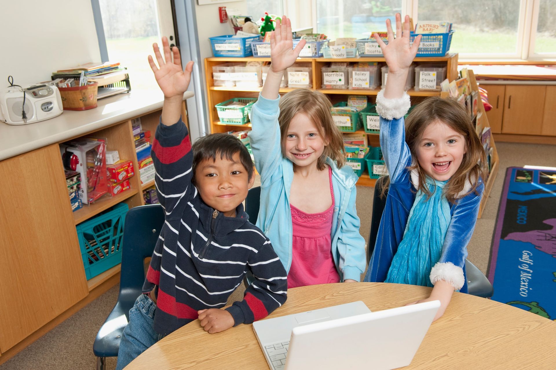 Three children smiling and raising hands at a classroom table with a laptop