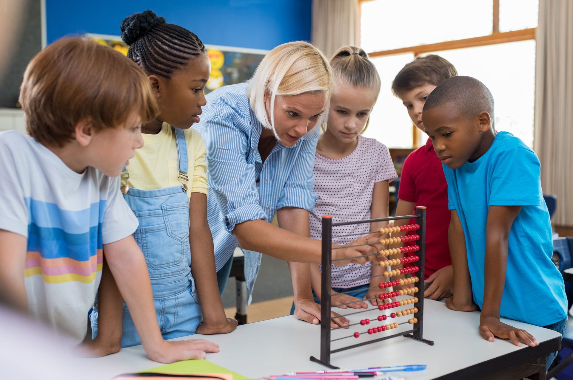 Children gathered around a classroom abacus, working together with a teacher at a table