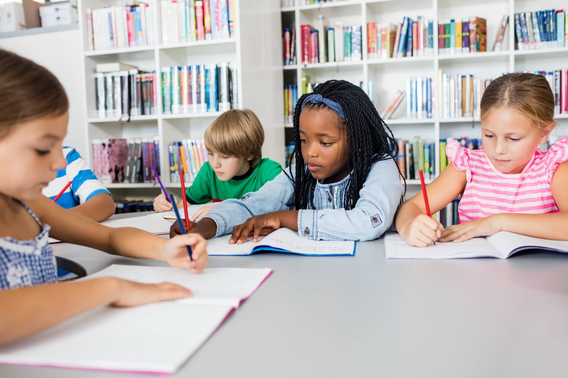 Children writing at a table in a library, surrounded by shelves of books