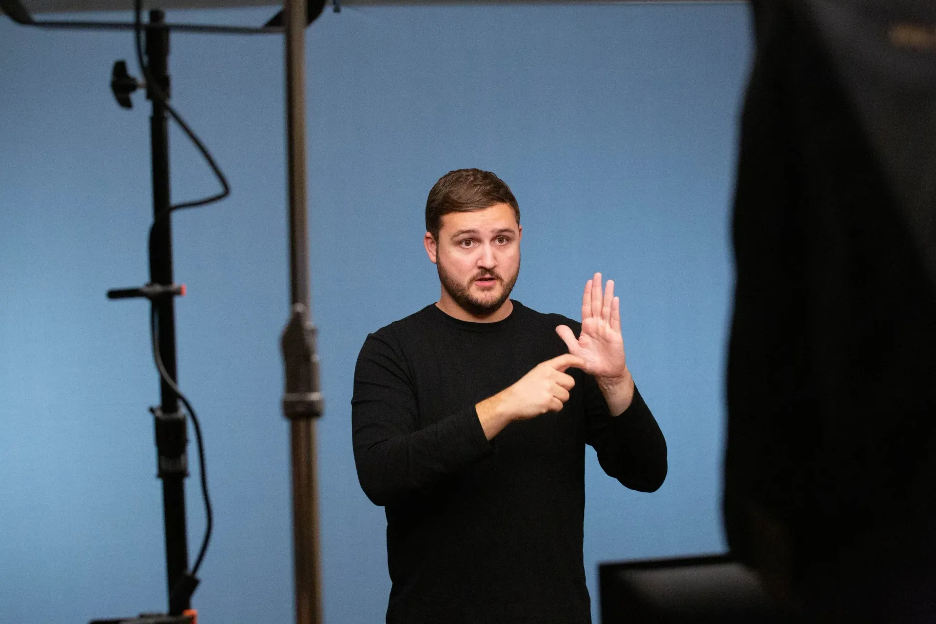Man in black shirt signing with hands in front of a blue backdrop and studio light stands