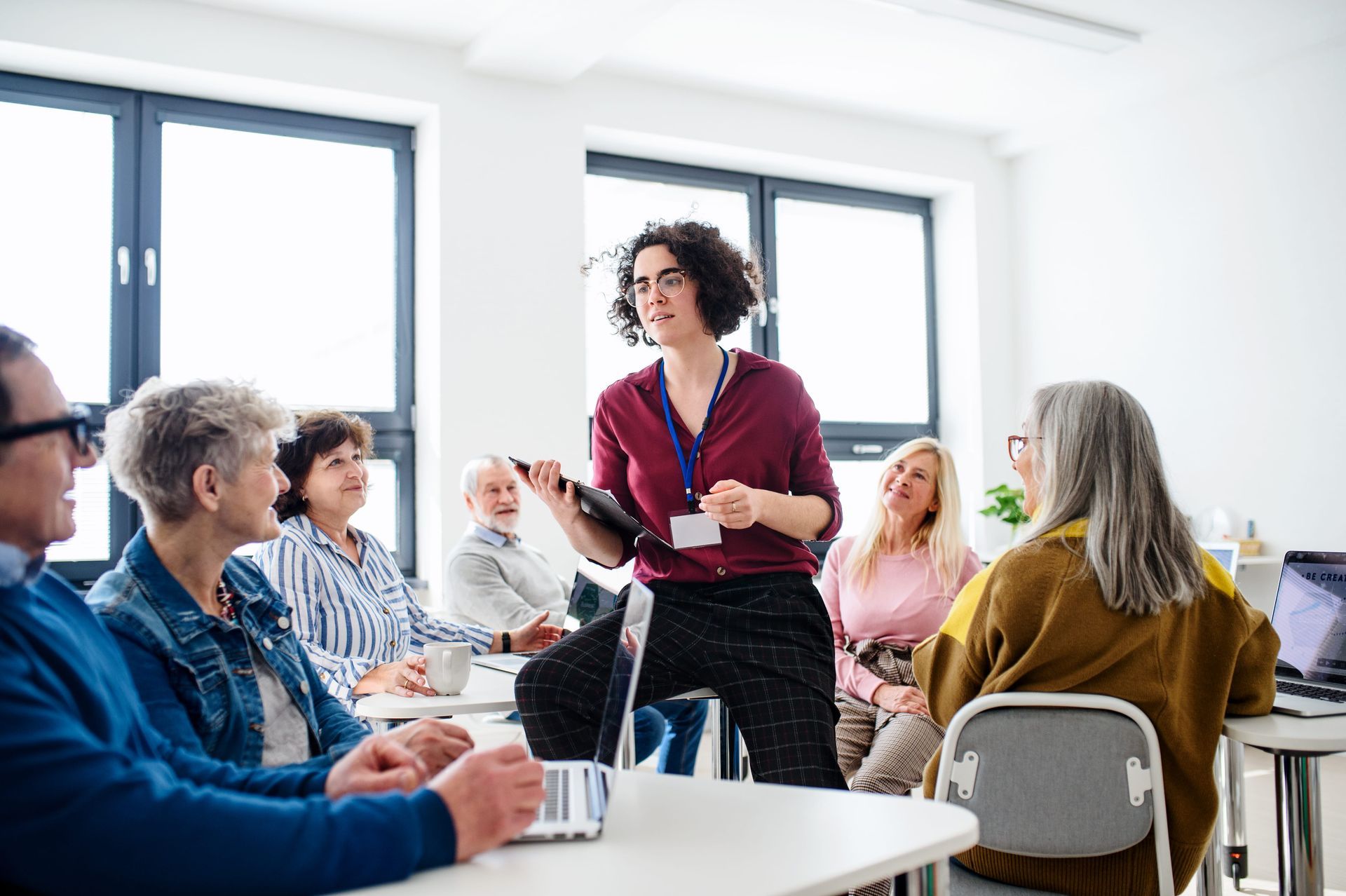 Presenter speaking to seated group in a bright meeting room, holding notes and smiling.