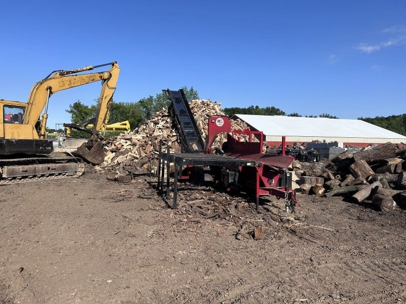 Excavator loading conveyor with wood debris at firewood processing site