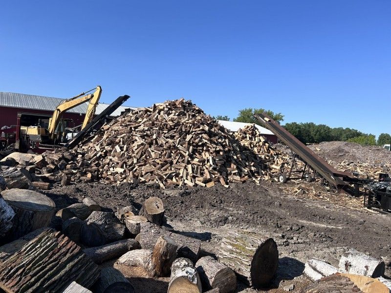 Excavator moving material next to firewood processing pile at tree service yard