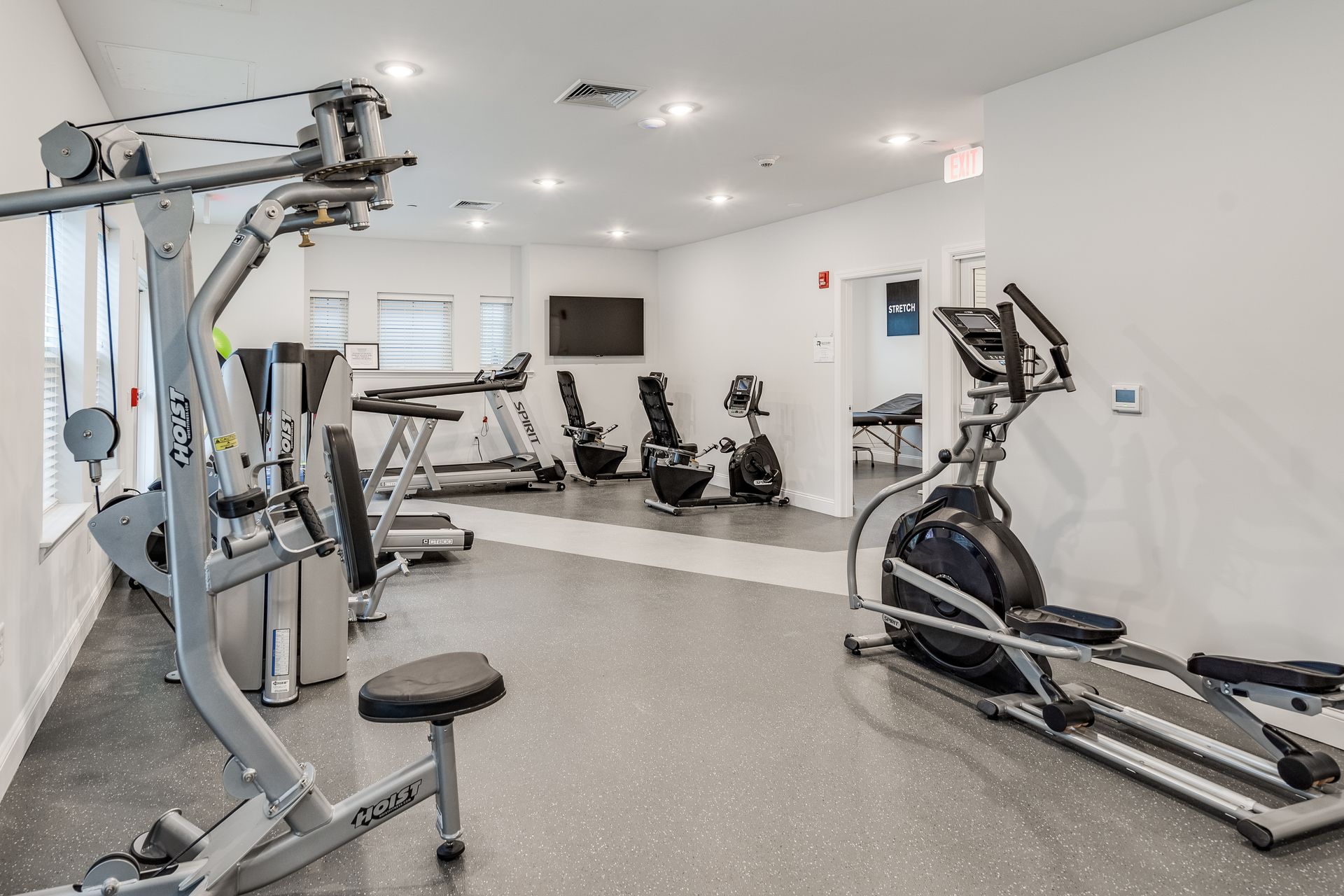 Gym with exercise equipment. Elliptical, treadmill, stationary bikes, weight machine. White walls, gray floor.