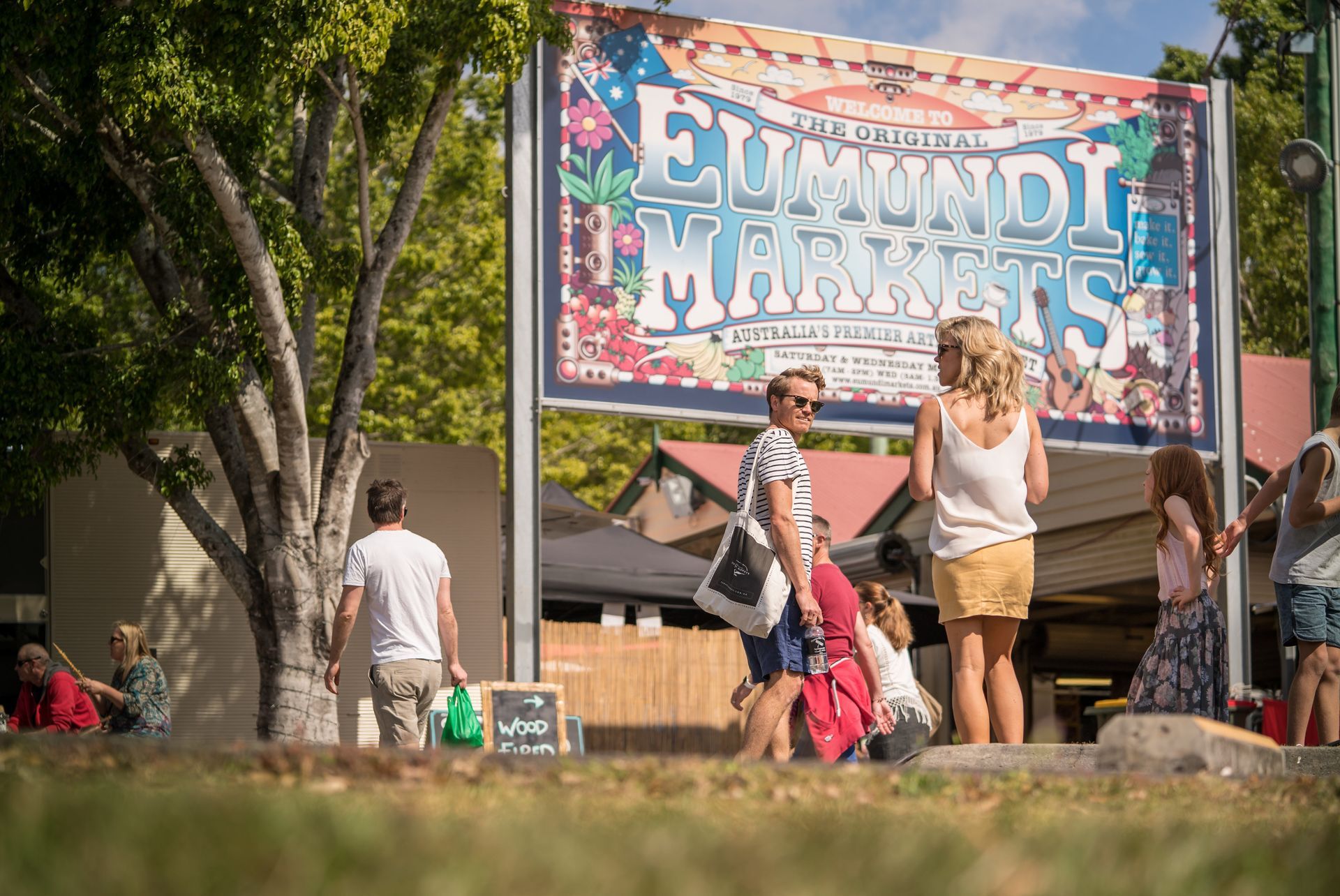 People at the Eumundi Markets entrance. Sign above, various people milling about, sunny day.