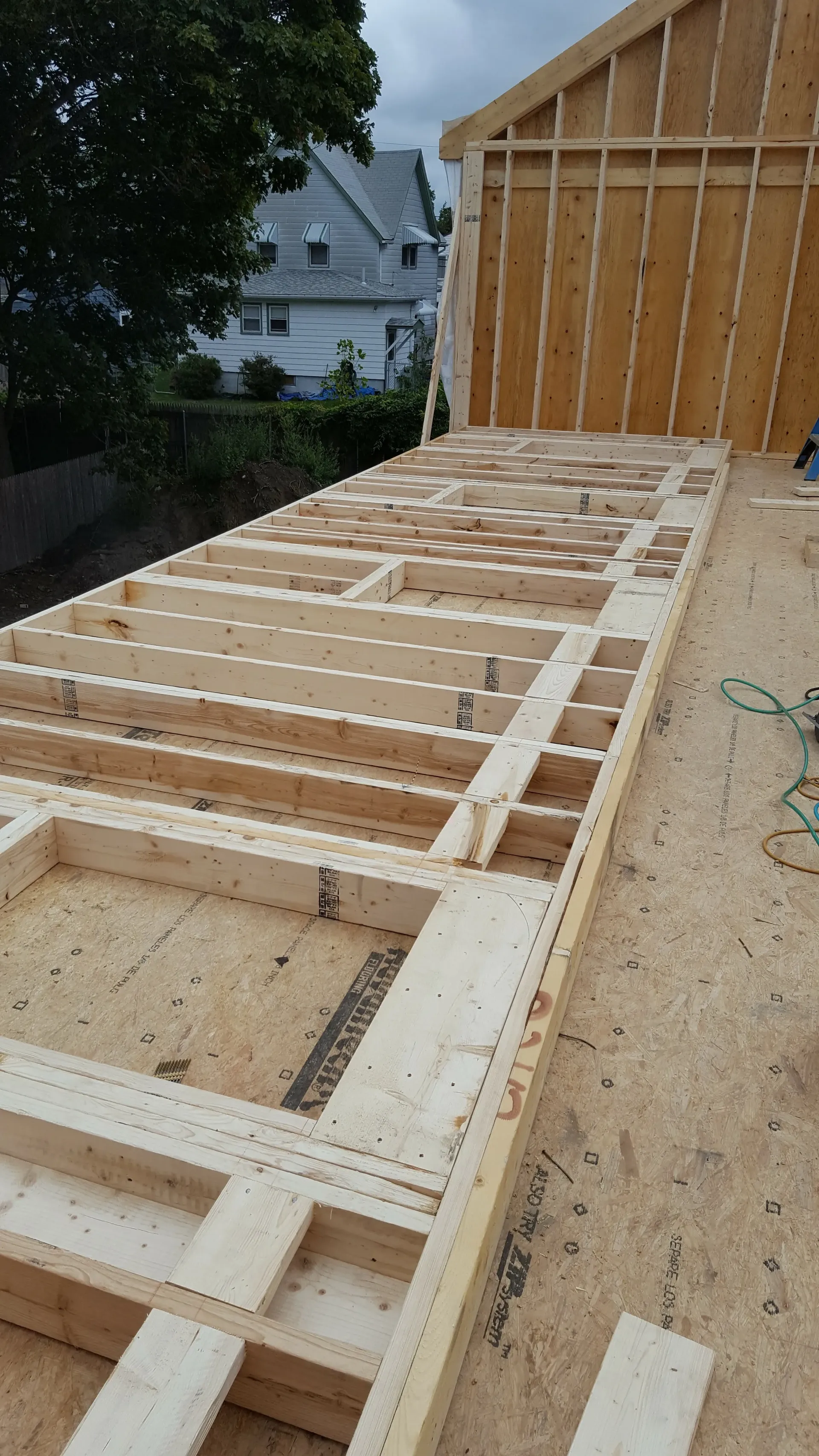 Wood-framed roof deck under construction beside a house, with exposed rafters and plywood sheathing