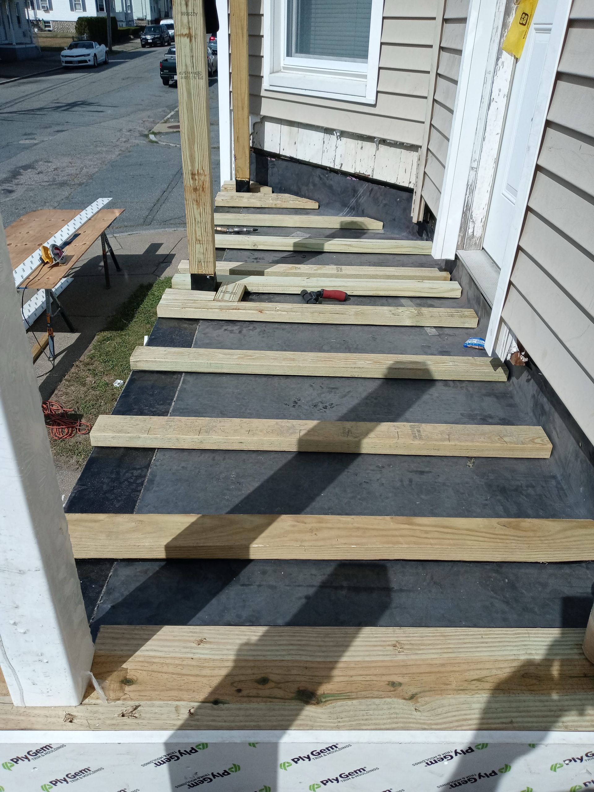 Wooden porch under construction with exposed joists, black underlayment, and scattered tools by the house entrance