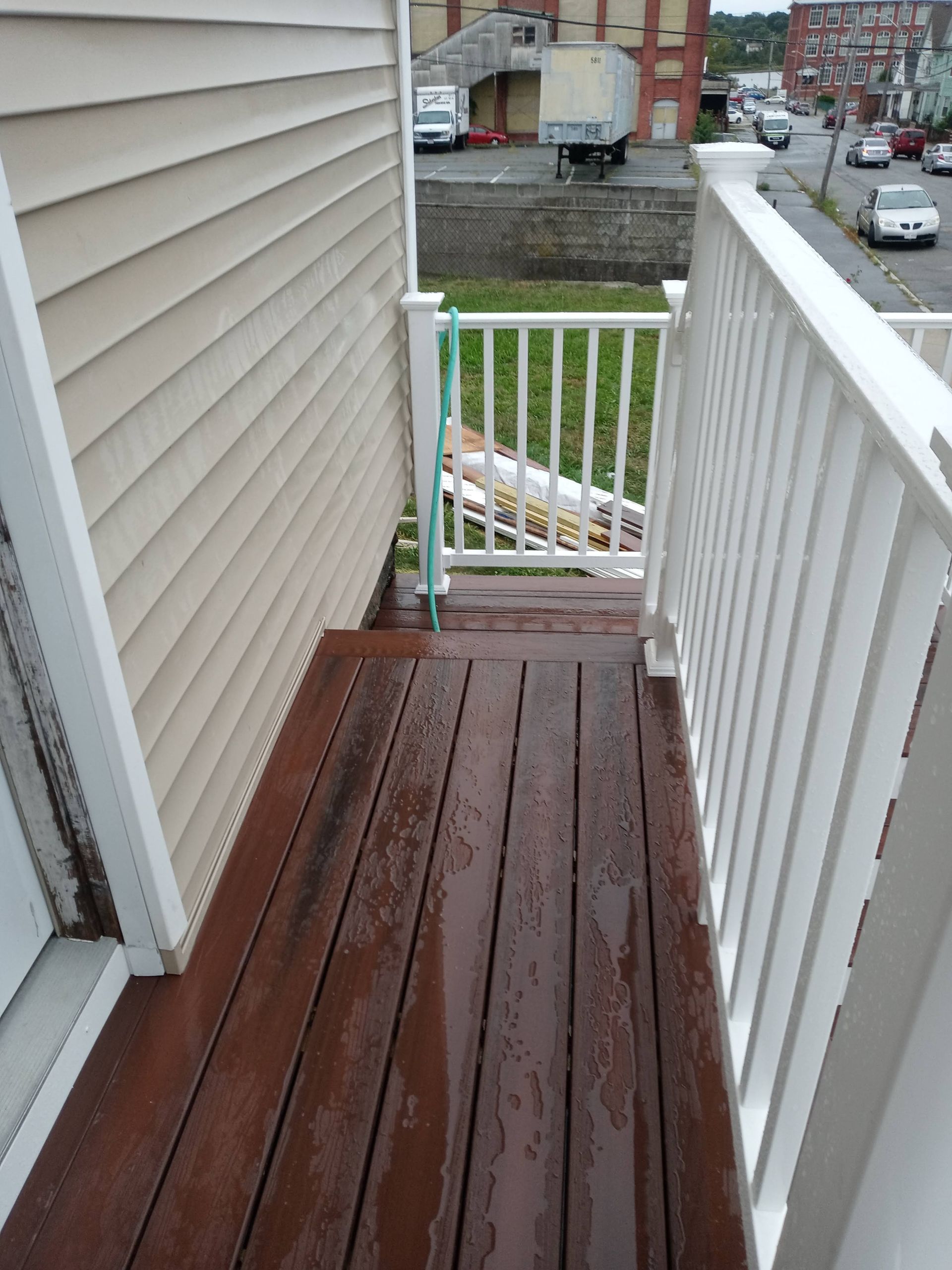 Narrow wooden porch with white railing, wet red floorboards, and a gated landing overlooking a street.