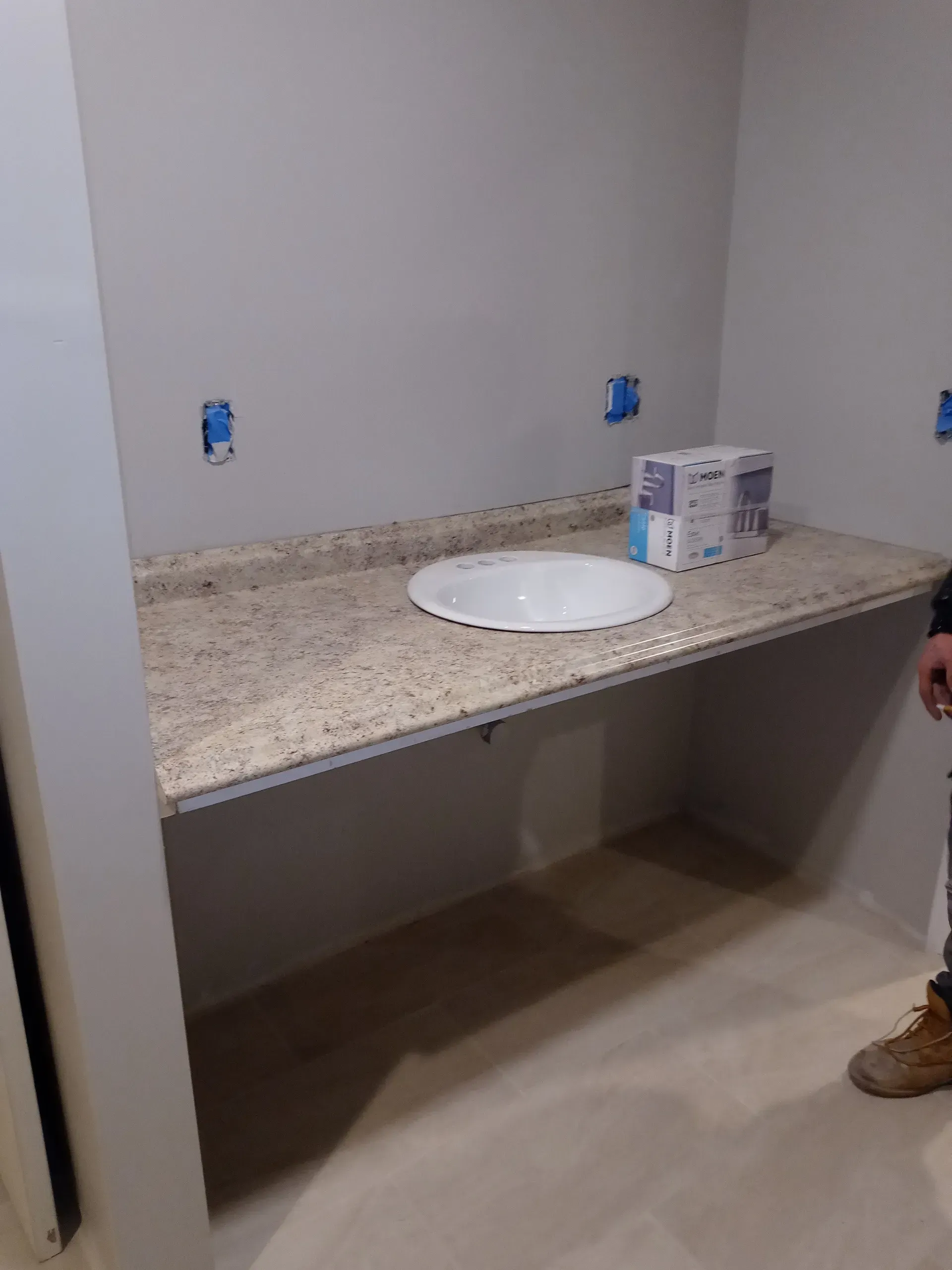 Unfinished bathroom vanity with granite countertop, white sink, exposed outlets, and a cardboard box on the counter.
