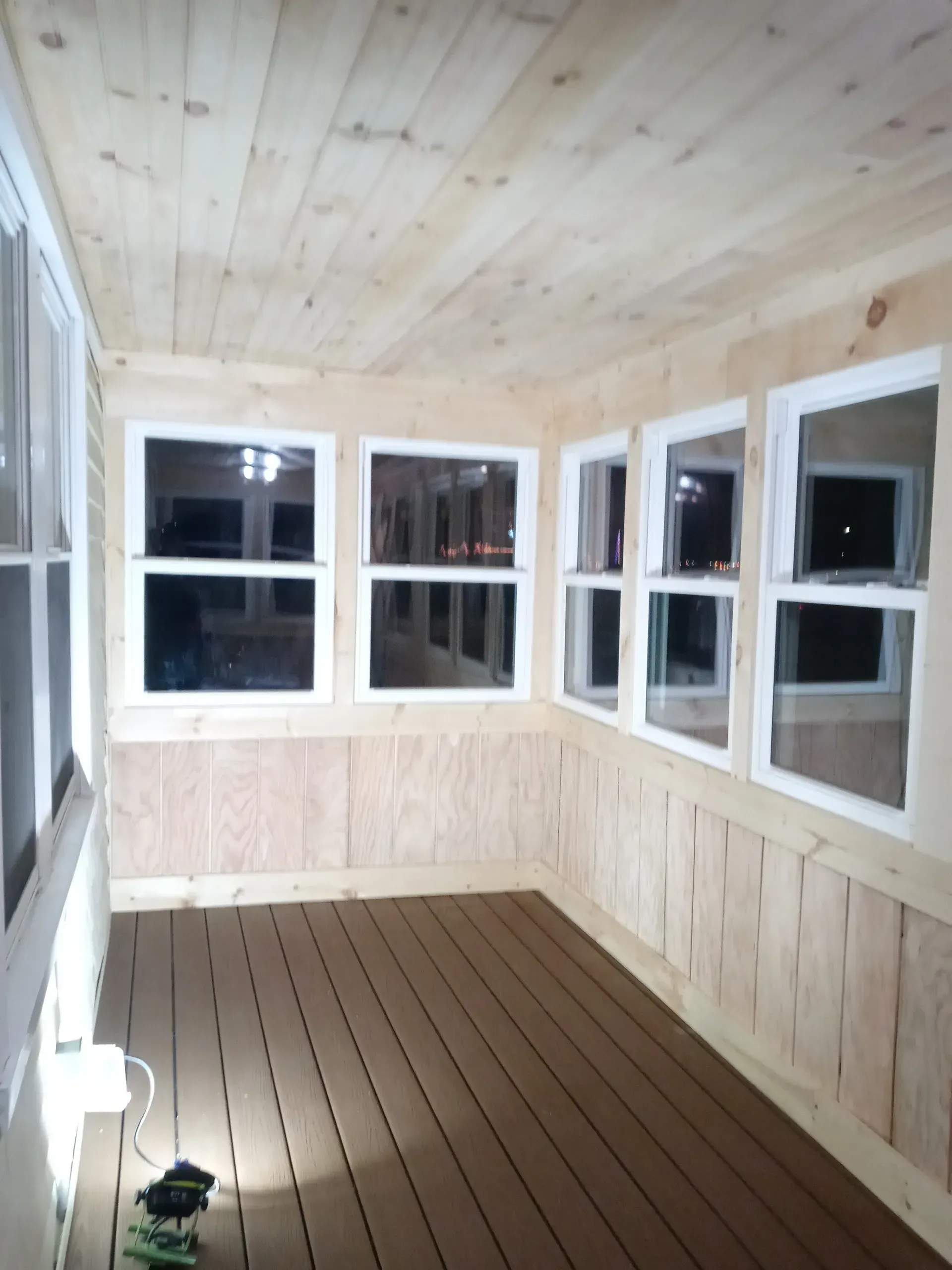 Small enclosed porch with light wood paneling, multiple windows, and a brown plank floor.
