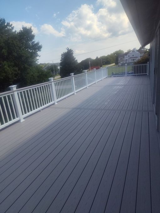 Empty wooden deck with white railing beside trees and a cloudy sky
